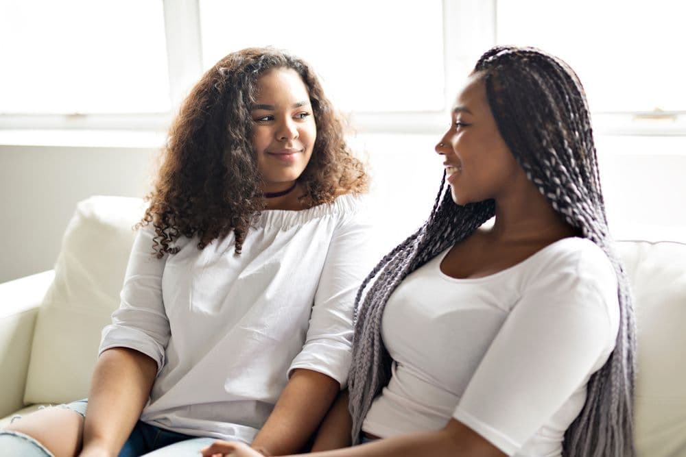 Two young women sit together on a sofa, talking