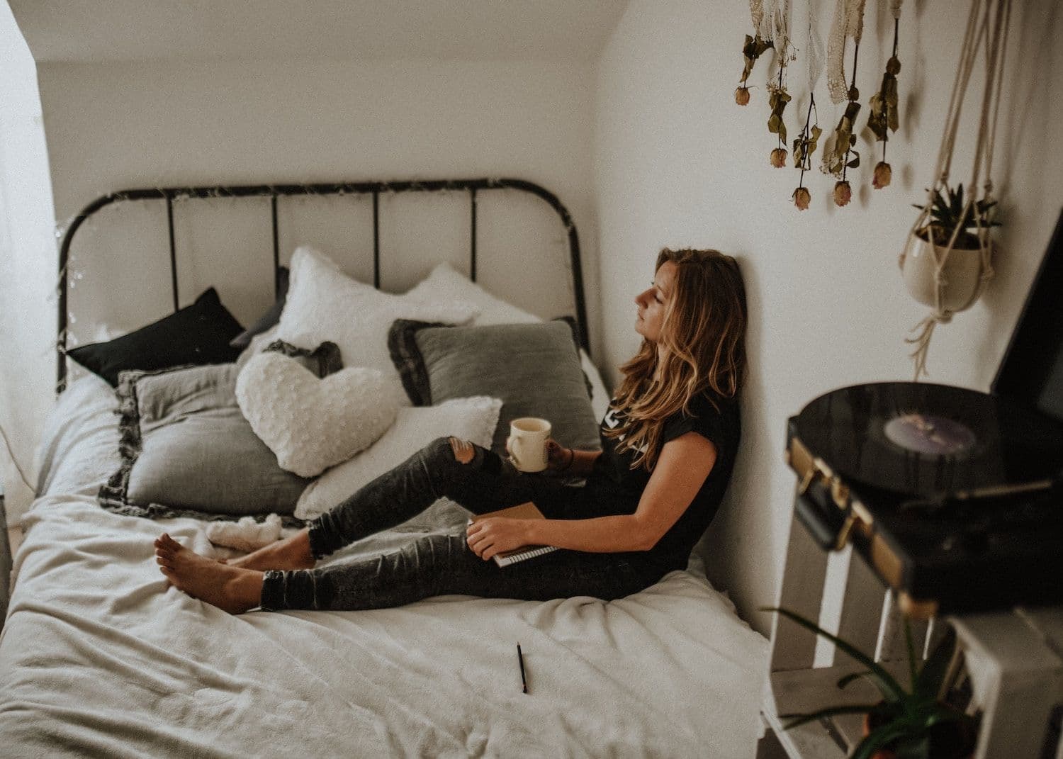 Woman sat on bed leaning against wall holding a mug and notebook. 