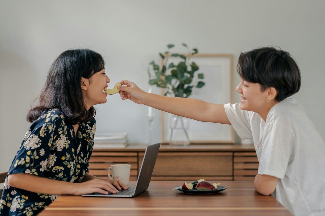 Two people are sat at a table, one is feeding the other apples as they work on a laptop.
