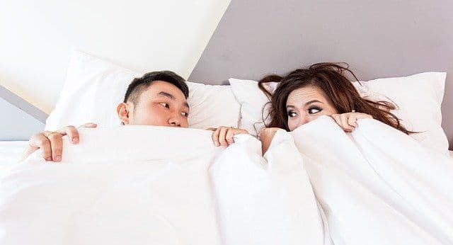 A couple look at each other nervously, peeking out from under a duvet