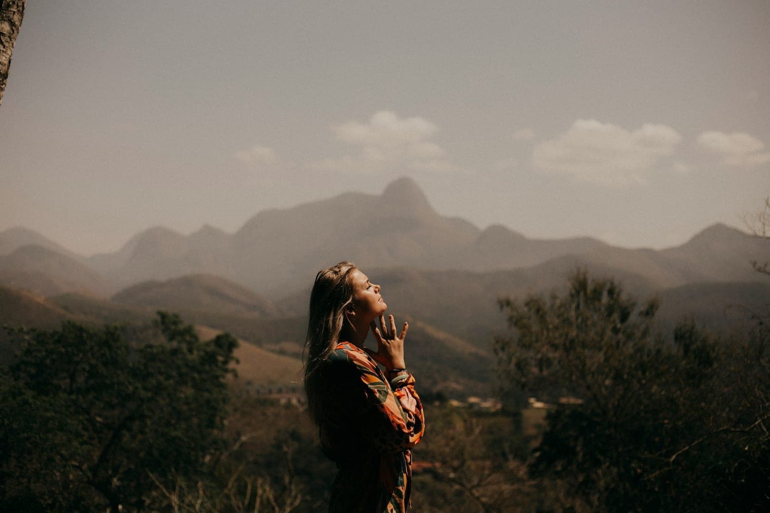 Woman stood in the mountains looking up with her hands under her chin in a praying pose.