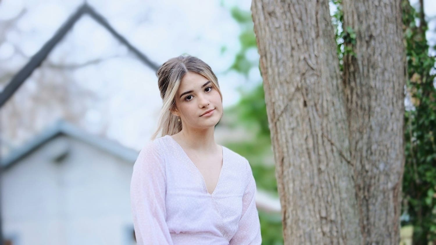 Image of a young girl stood by a tree, with a neutral expression.